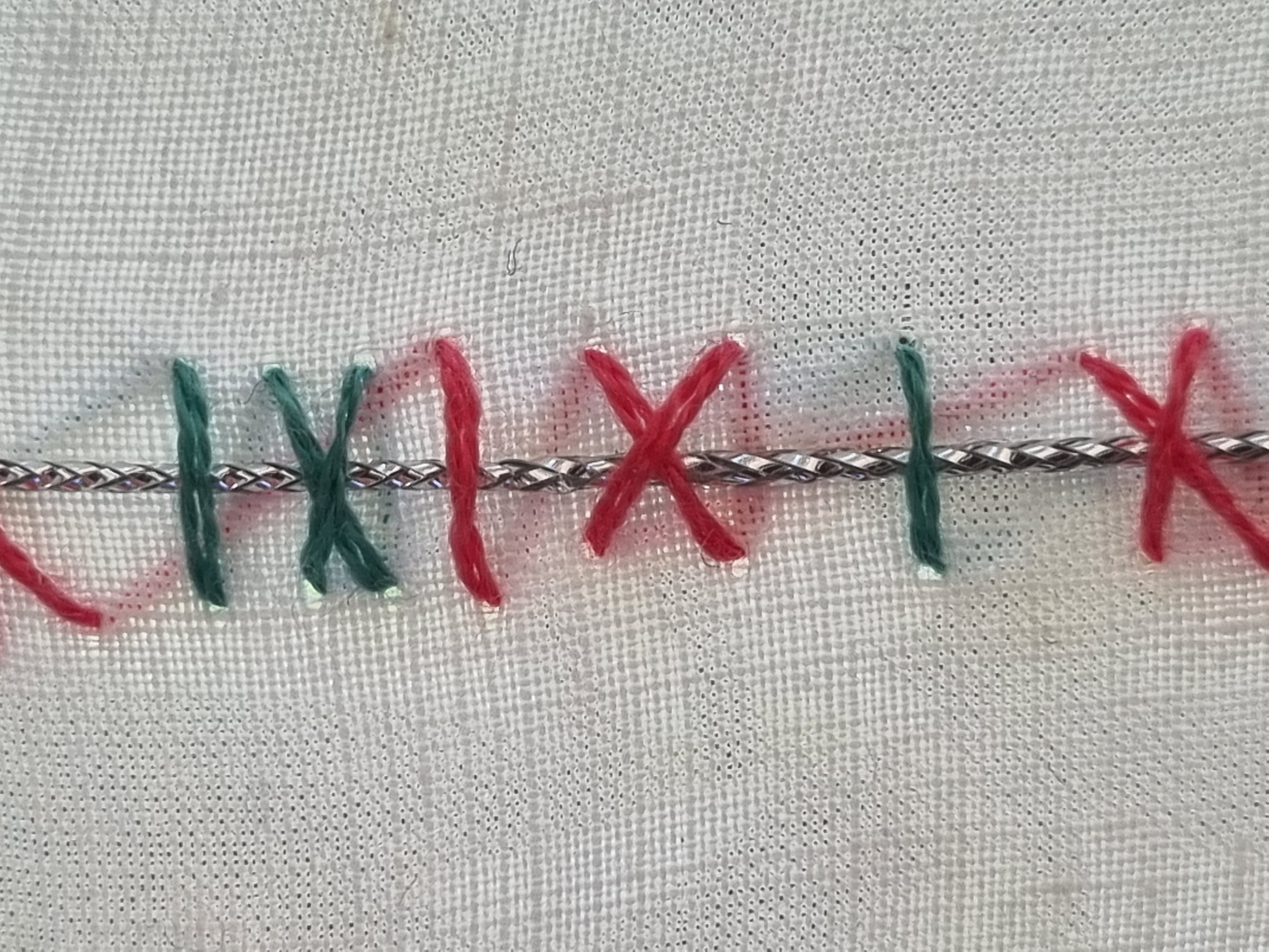 Extreme close-up of the sampler: a braided silver laid thread crosses open-weave white linen, held by green straight and crossed stitches and magenta straight, slanted and crossed stitches.