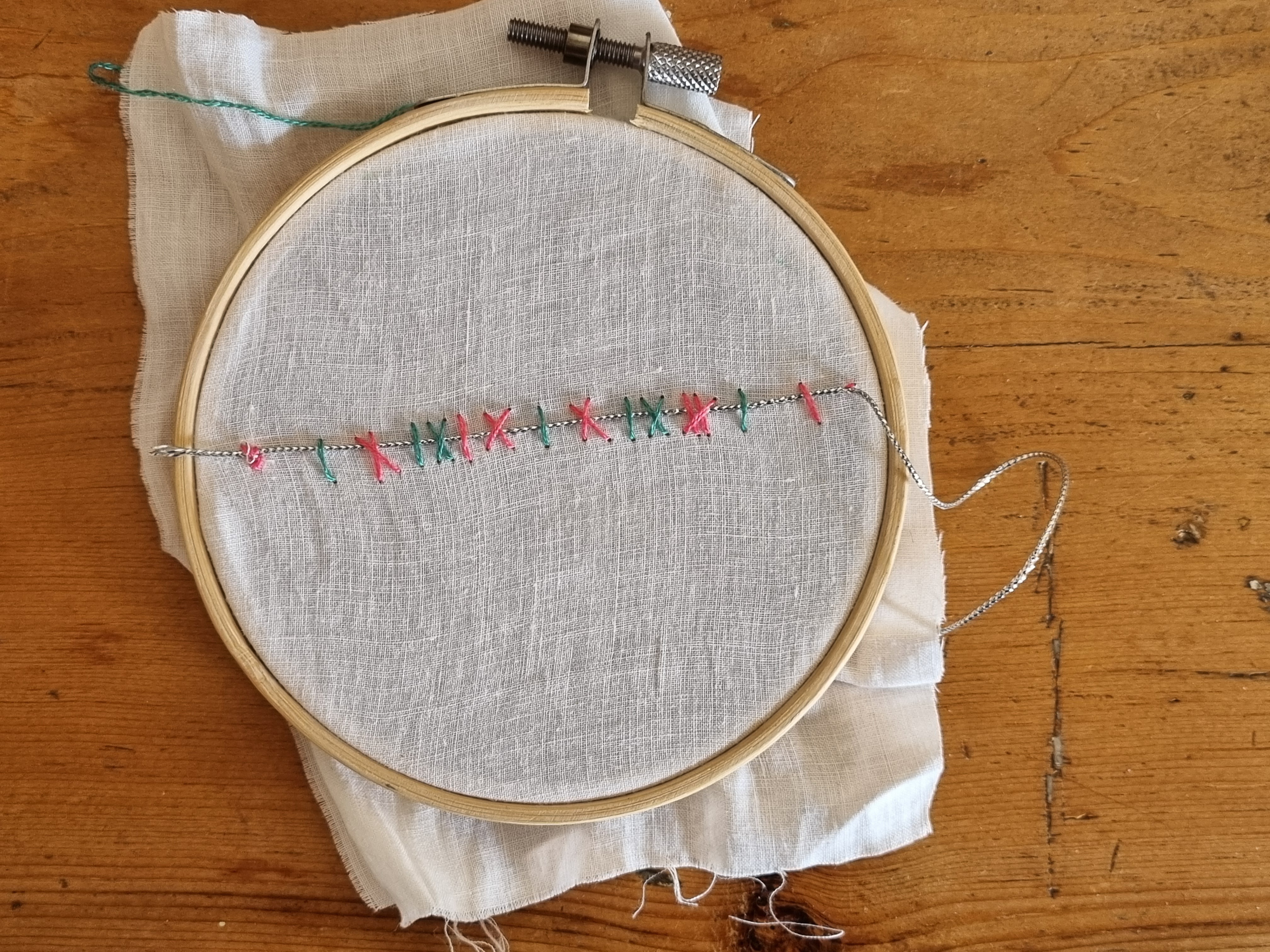 A complete embroidery sampler in a wooden hoop on a wooden table. A silver thread crosses white linen, held by alternating magenta and green stitches, with loose thread continuing beyond the hoop.